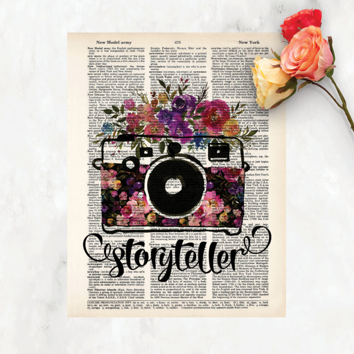 storyteller with watercolor camera and flowers in the body of the camera and on top in shades of pinks, purples, yellow, and blue with greenery printed on a dictionary page