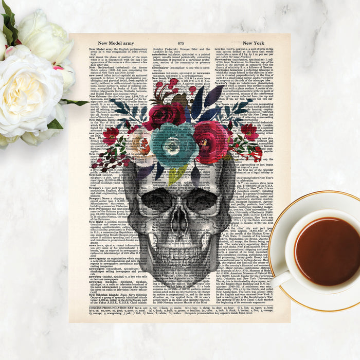 pencil sketch of a skull looking straight on topped with watercolor flowers in plue, pale pink, red, deep red and muted greenery on top printed on a dictionary page