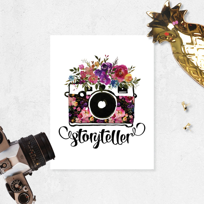 storyteller with watercolor camera and flowers in the body of the camera and on top in shades of pinks, purples, yellow, and blue with greenery printed on matte white paper
